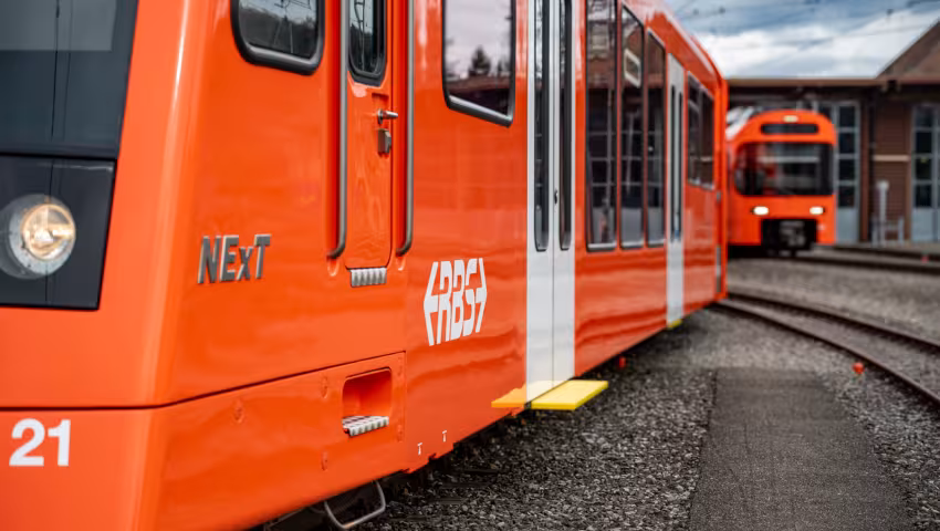 Orangefarbener modernisierter NExT‑Zug der RBS auf Abstellgleisen, seitliche Ansicht mit Türen und zweitem Zug im Hintergrund.