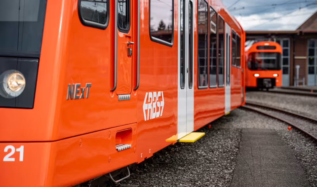 Orangefarbener modernisierter NExT‑Zug der RBS auf Abstellgleisen, seitliche Ansicht mit Türen und zweitem Zug im Hintergrund.