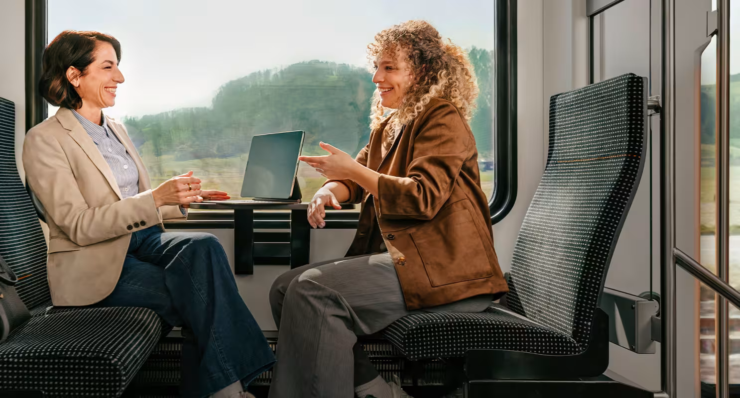 Fahrgäste im RBS-Zug sitzen sich im Abteil gegenüber und führen ein Gespräch, Landschaft durch das Fenster sichtbar.