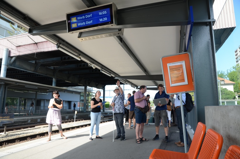 Auf dem Perron stehen viele Testpersonen, welche die Abfahrtsmonitore begutachten