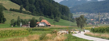 worbletalwärts - ein Blick in das Worblental. Links steht ein Bauernhaus im Grünen, rechts ein schotterweg, auf dem eine frau mit Hund und Kind auf einem LikeaBike stehen.